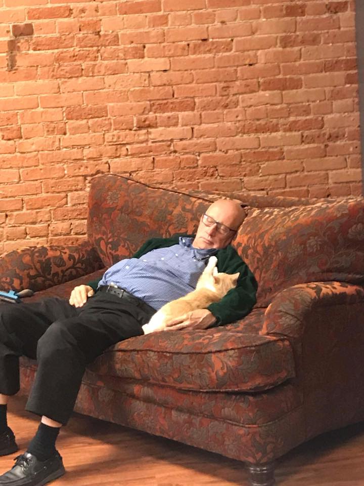 A man sleeps on a sofa with a cat curled up on his arm.