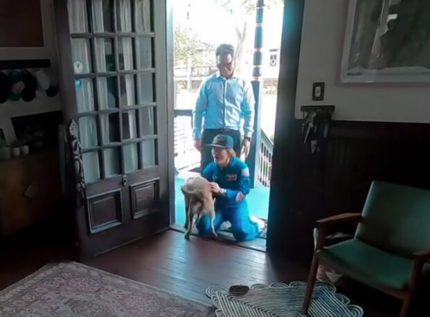 A woman kneels in the doorway and embraces a dog. A man is standing behind them.