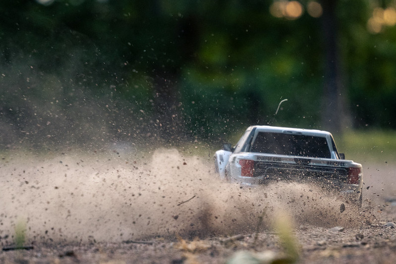 a radio-controlled toy car drives across a dirt field, kicking up dust and pebbles