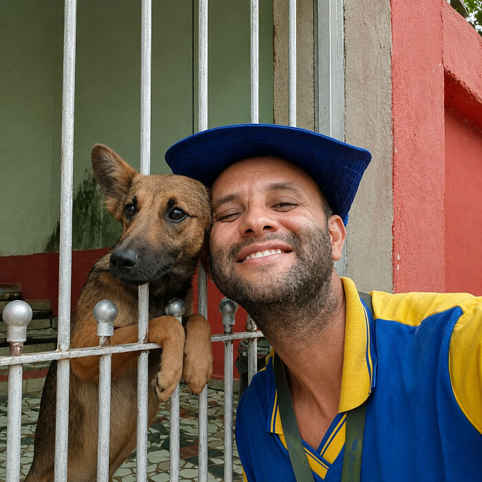 A dog pokes its head through a metal gate to press against a smiling man on the other side.