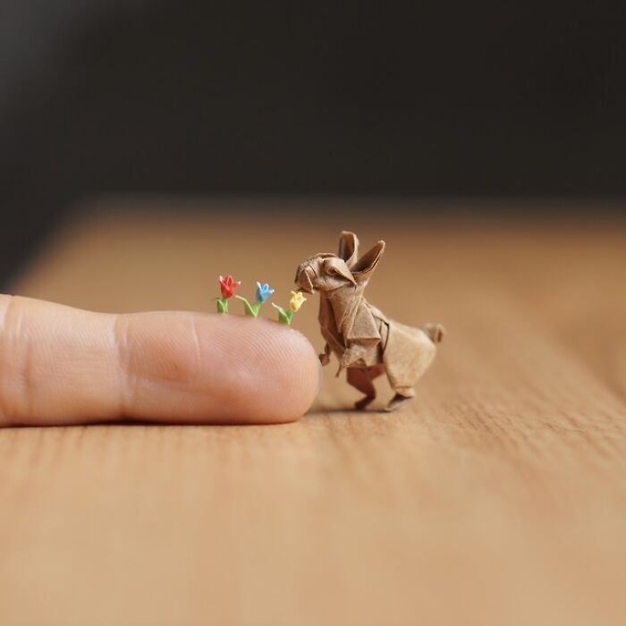 A human finger pointing to very small sculpture of a rabbit. Three miniature flowers are lined up on the finger and the rabbit is facing them. 