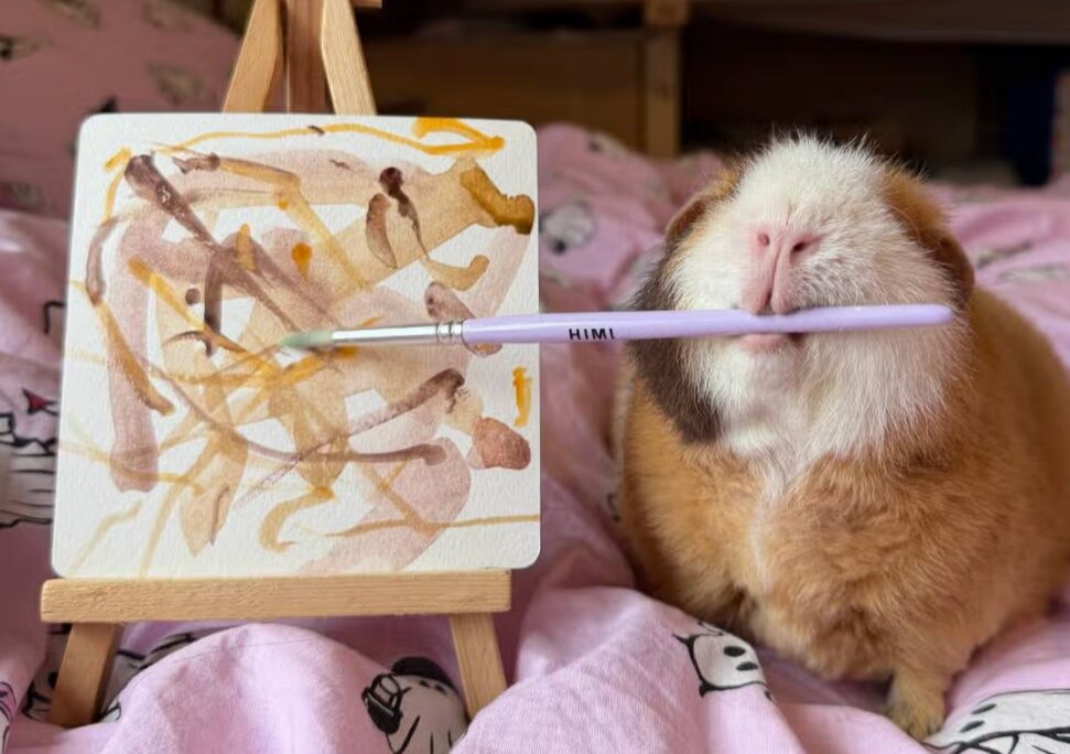 A guinea pig holds a paintbrush in its mouth and paints on a small canvas