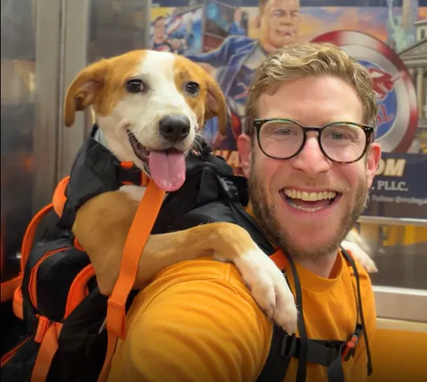 A smiling dog rides in a backpack worn by a smiling man